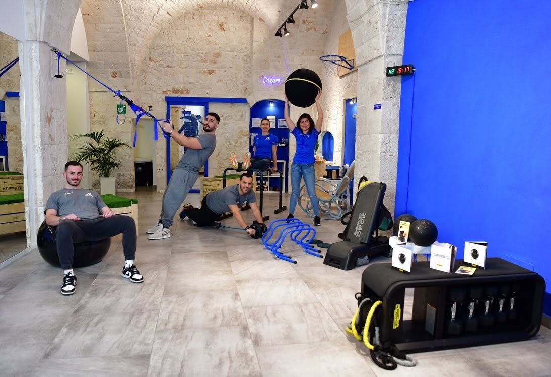 Cinque istruttori di AD Performance con fitball, cinghie e ostacoli blu in una sala con volte in pietra e neon «Dream