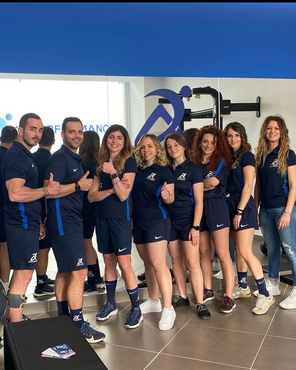 Team di AD Performance in polo e pantaloncini blu con pollici alzati davanti al logo, sala pesi con pavimento grigio